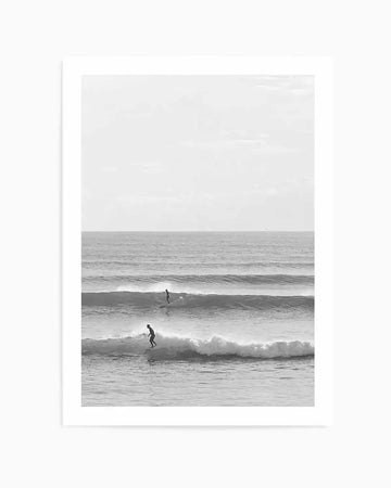 Framed black and white photograph of a coastal beach scene with a white frame and white border. The composition features two surfers riding waves in the ocean under an overcast sky.