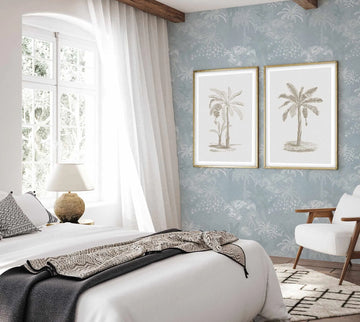 Lifestyle shot of a bedroom featuring Hotel Bahamas In Ocean wallpaper. The wallpaper has a repeating pattern of white and light grey palm trees and tropical foliage against a soft blue background. Two framed art prints featuring sepia-toned illustrations of palm trees are displayed on the wallpaper above a white armchair with wooden arms. A bed with white linen, patterned throw, and decorative pillows is visible in the foreground, with a large window and sheer white curtains to the left.