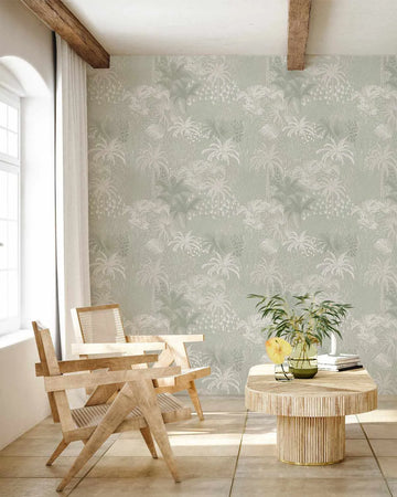 Lifestyle shot of a living room featuring Hotel Bahamas in Sage wallpaper. The wallpaper has a repeating pattern of palm trees and tropical foliage in varying shades of sage green and off-white on a light sage green background, with a textured, hand-drawn quality. The room includes two light wood chairs with woven cane seats, a round light wood coffee table with vertical slat detailing, and a large window with white curtains.