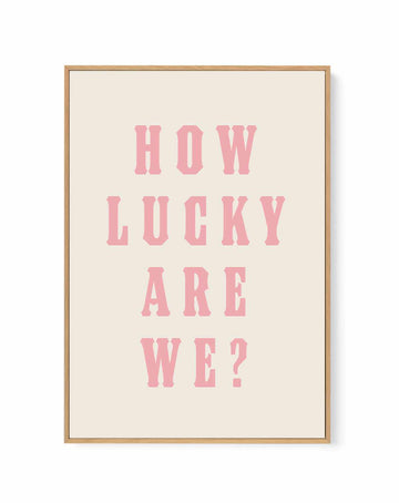 Framed canvas with bold pink serif text 'How Lucky Are We?' on cream ground, natural wood float frame