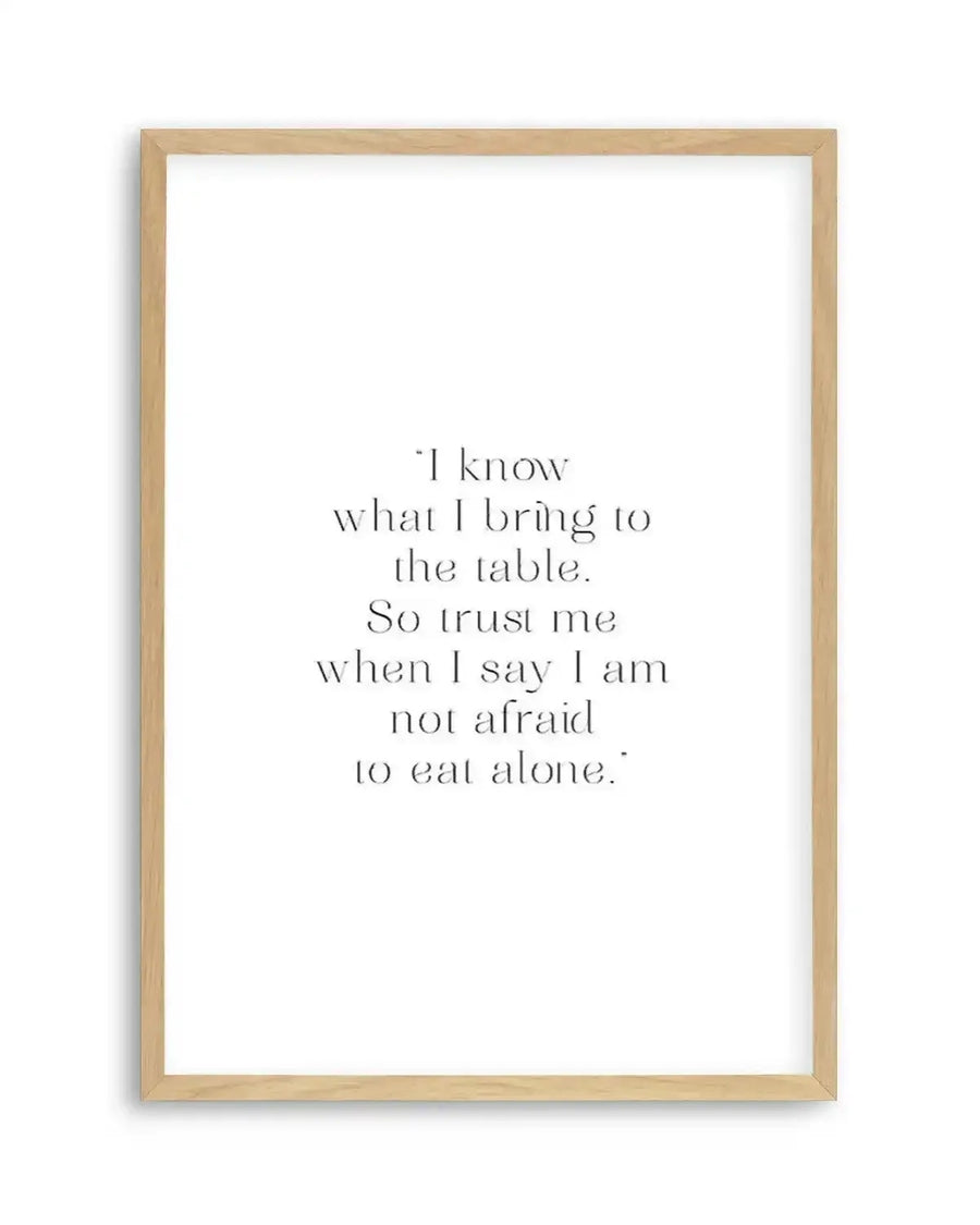 Art print featuring a motivational quote in black serif font on a crisp white background, displayed in a natural oak frame with white border. The quote reads: "I know what I bring to the table. So trust me when I say I am not afraid to eat alone."