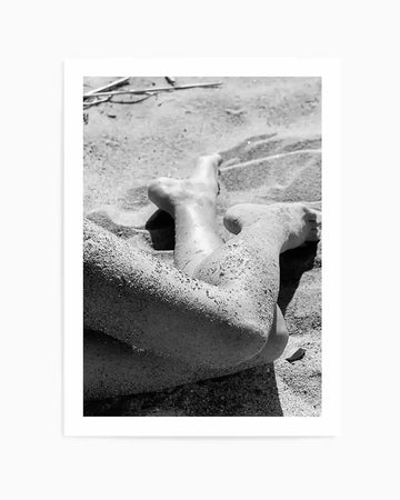 Framed black and white photograph of a person's legs and feet partially covered in sand, lying on a sandy beach. The composition captures the texture of the sand and the subtle curves of the body, with a natural wood frame and white border.