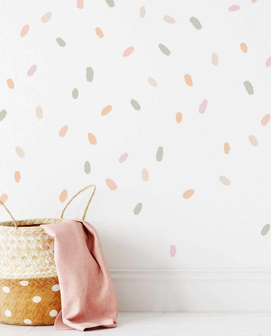 Wall Decal:
"Peel and stick wall decals featuring a scattered pattern of irregular, hand-painted brushstroke dots in a palette of pastel pink, peach, sage green, and cream on a white background. The dots vary slightly in size and shape, creating a playful, confetti-like effect. A woven basket with a pink linen cloth is visible in the bottom left corner."