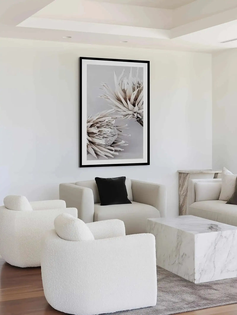 Framed art print displayed on a white wall above cream-colored seating. The print shows a close-up photograph of two protea flower heads in muted greige and cream tones against a soft gray background. The flowers feature layered, pointed petals with fine botanical detail. Black frame with white border. Room features modern minimalist design with curved cream upholstered chairs, white marble side table, and warm wood flooring.