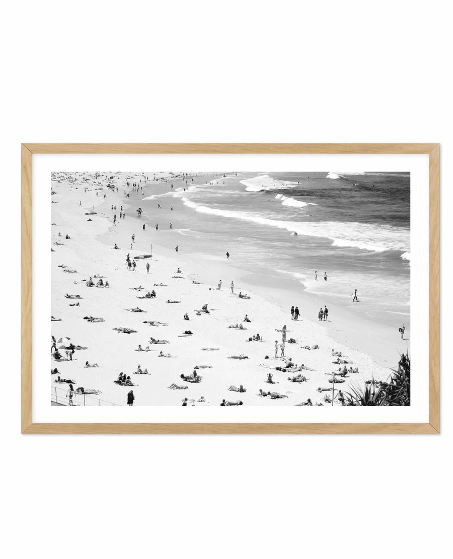 Black and white aerial photograph of Bondi Beach showing a crowded sandy beach with numerous beachgoers scattered across the sand, ocean waves with white foam in the background, and people swimming and wading in the water. The photograph is printed on white paper and framed in a natural light wood frame with a white border, creating a clean, minimalist presentation of this iconic Australian beach scene.