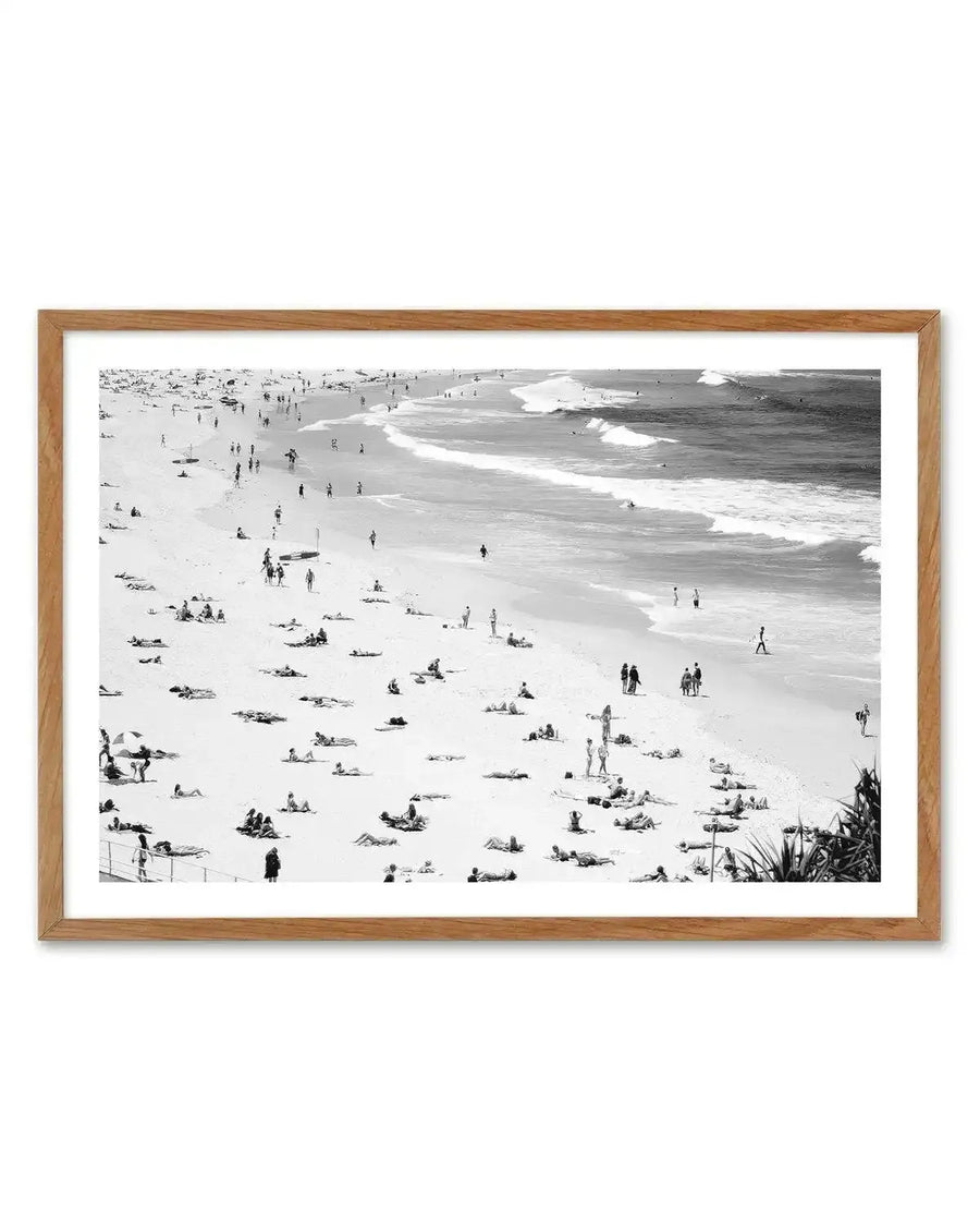 Black and white aerial photograph of Bondi Beach framed in light natural wood. The image captures a crowded beach scene from above, showing scattered groups of beachgoers on pale sand, swimmers in the ocean with visible white-capped waves, and small clusters of people throughout the composition. The photograph has high contrast, crisp detail, and a documentary photography style. A white border surrounds the image within the wooden frame.