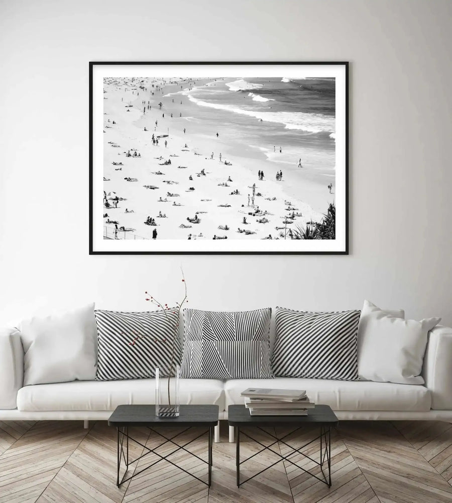 Black and white aerial photograph of Bondi Beach displayed in a black frame with white border, hanging above a cream linen sofa. The photograph captures a bird's-eye view of the sandy beach scattered with beachgoers, umbrellas, and swimmers in the ocean with rolling waves. The monochrome image shows fine detail and contrast, printed on matte paper. The framed print is styled in a minimalist living room with striped accent pillows and a dark metal coffee table below.