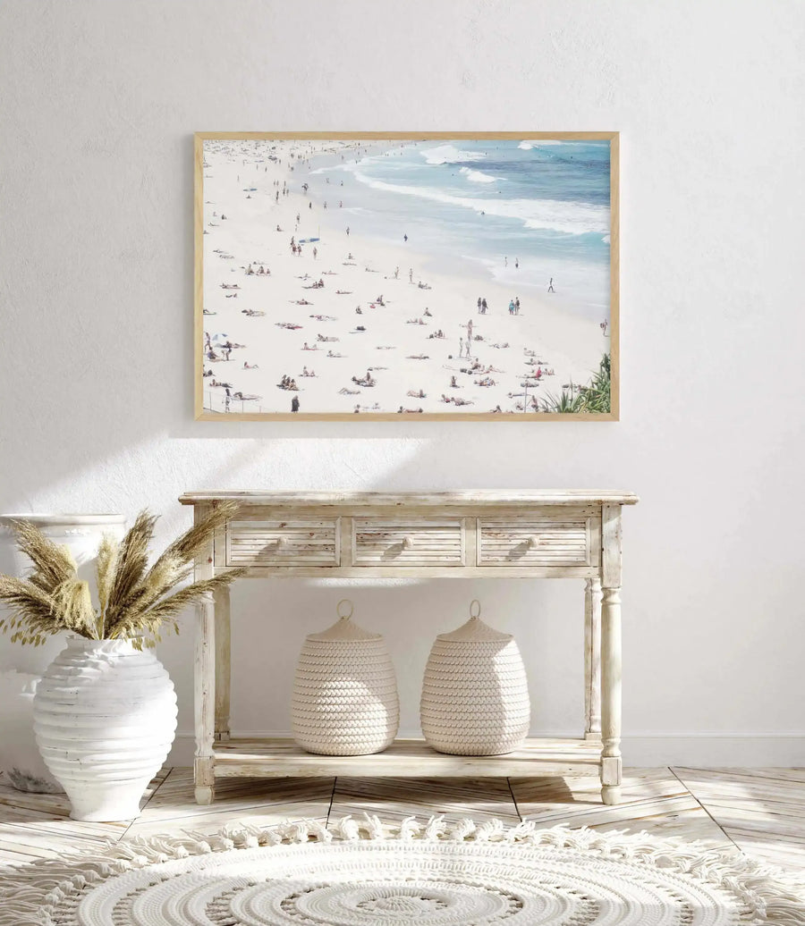 Lifestyle shot of an art print displayed above a rustic wooden console table in a bright room. The art print features an aerial view of a crowded beach with many small figures sunbathing and swimming in the light blue ocean, with a natural wood frame and white border. The console table has three drawers and a lower shelf, with two woven baskets on the shelf and a large white vase with dried pampas grass on the floor next to it. A round, white textured rug is on the light wood floor.