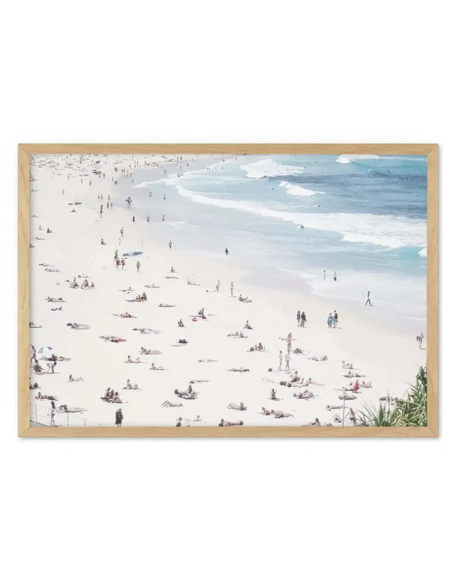 Framed art print featuring an aerial perspective of a crowded beach scene with a natural wood frame and white border. The composition captures numerous tiny figures sunbathing and walking on the light beige sand, with gentle turquoise waves breaking on the shore under a bright sky.