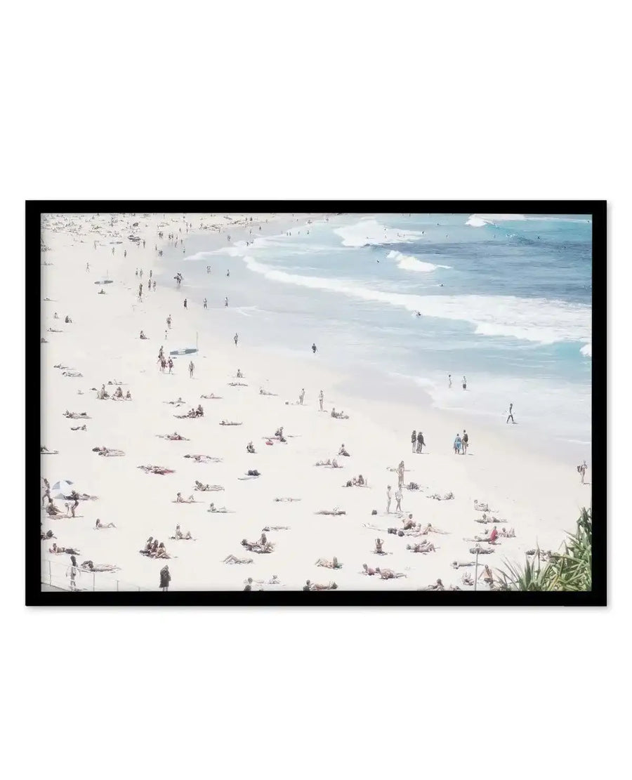 Framed photograph of a crowded beach scene with a black frame and white border. The composition features a wide expanse of light sandy beach with numerous people sunbathing and walking, leading to the light blue ocean with white waves breaking.