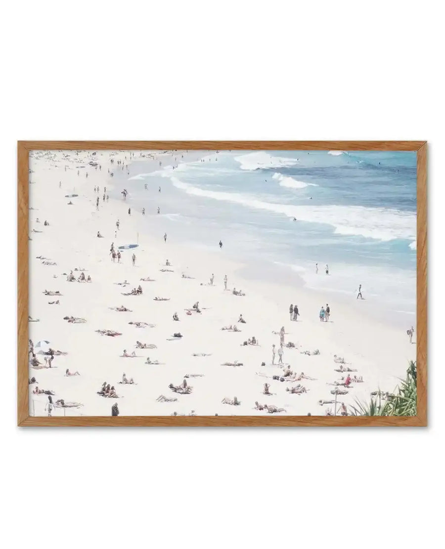 Framed art print of an aerial photography shot of a crowded white sand beach with people sunbathing and walking along the shore, and gentle blue waves breaking in the ocean, displayed in a natural oak frame with white border.
