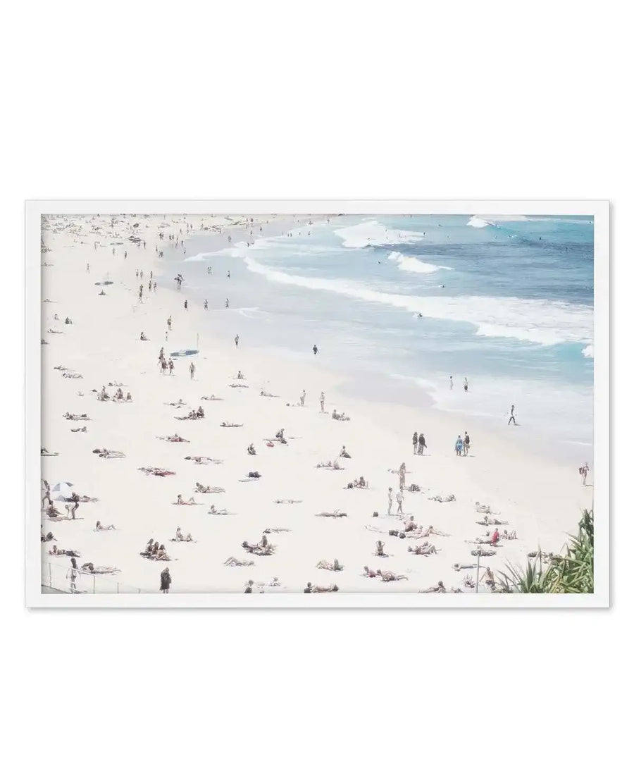 Framed art print featuring a high-angle photograph of a crowded beach with many people sunbathing on the light beige sand and swimming in the pale blue ocean with white waves. The print is displayed in a white frame with a white border.