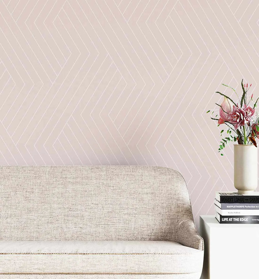 "Blush pink wallpaper with a repeating geometric pattern of thin white lines in a chevron or herringbone style, displayed in a minimalist living room. A light beige textured sofa is in the foreground, and a white side table with a stack of books and a vase of pink flowers is to the right."