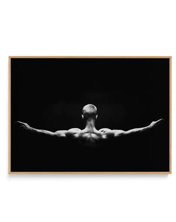 Black and white photograph of a muscular man viewed from behind with arms outstretched to the sides in a triumphant pose against a solid black background. The image is printed on canvas and framed with a natural wood-tone frame. High-contrast monochromatic composition emphasizing the figure's physique and expansive arm position. Landscape orientation.