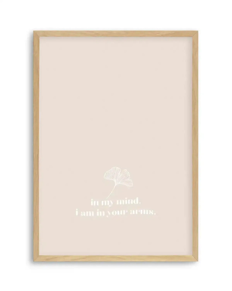 Framed art print with natural wood frame featuring a cream-colored background. A delicate white line illustration of a ginkgo leaf is centered in the upper-middle portion. Below the leaf, white sans-serif text reads "in my mind, i am in your arms." The print has a minimalist aesthetic with subtle, gentle composition on what appears to be a slightly textured off-white surface with a thin white border around the edges.