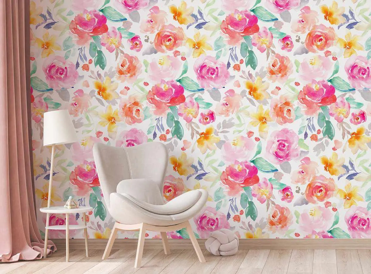 Lifestyle shot featuring a room with a wall covered in "In a Daydream Wallpaper." The wallpaper features a repeating pattern of hand-painted watercolor flowers in shades of fuchsia, blush pink, coral, and bright yellow, with soft green and muted purple leaves on a white background. The design has a loose, organic watercolor quality with visible brushstroke texture. A light-colored armchair, a small round side table with a lamp, and a pink curtain are visible in the foreground.