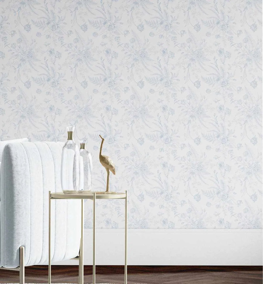 Lifestyle shot of a room with "In the Hamptons" wallpaper featuring a repeating pattern of delicate botanical illustrations in soft blue on a white background. The wallpaper shows clusters of various flowers and ferns with a hand-drawn, slightly faded aesthetic. A light-coloured, upholstered sofa is partially visible on the left, next to a small, round gold side table holding two clear glass bottles and a gold crane figurine. The room has dark wood flooring and white baseboards.