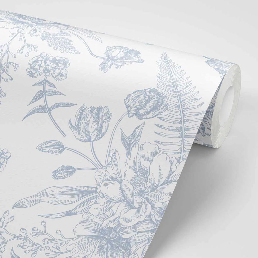 Peel and stick wallpaper featuring a repeating pattern of hand-drawn botanical illustrations in a soft, muted blue on a crisp white background. The design includes various flowers such as peonies, tulips, and smaller blossoms, along with ferns and other foliage, creating a classic garden aesthetic. The wallpaper is shown partially unrolled, revealing the pattern and texture of the paper.