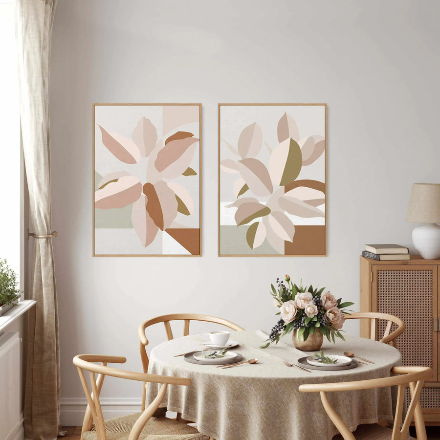 Two framed canvas art prints featuring abstract botanical designs, displayed side-by-side on a light grey wall in a dining room. Each print depicts overlapping, rounded leaf shapes in shades of dusty rose, blush pink, sage green, and warm brown, against a light cream background, with natural wood frames. The dining room includes a round table with a cream linen tablecloth, light wood chairs, a vase of pink peonies and dark berries, and a wooden cabinet with cane detailing.