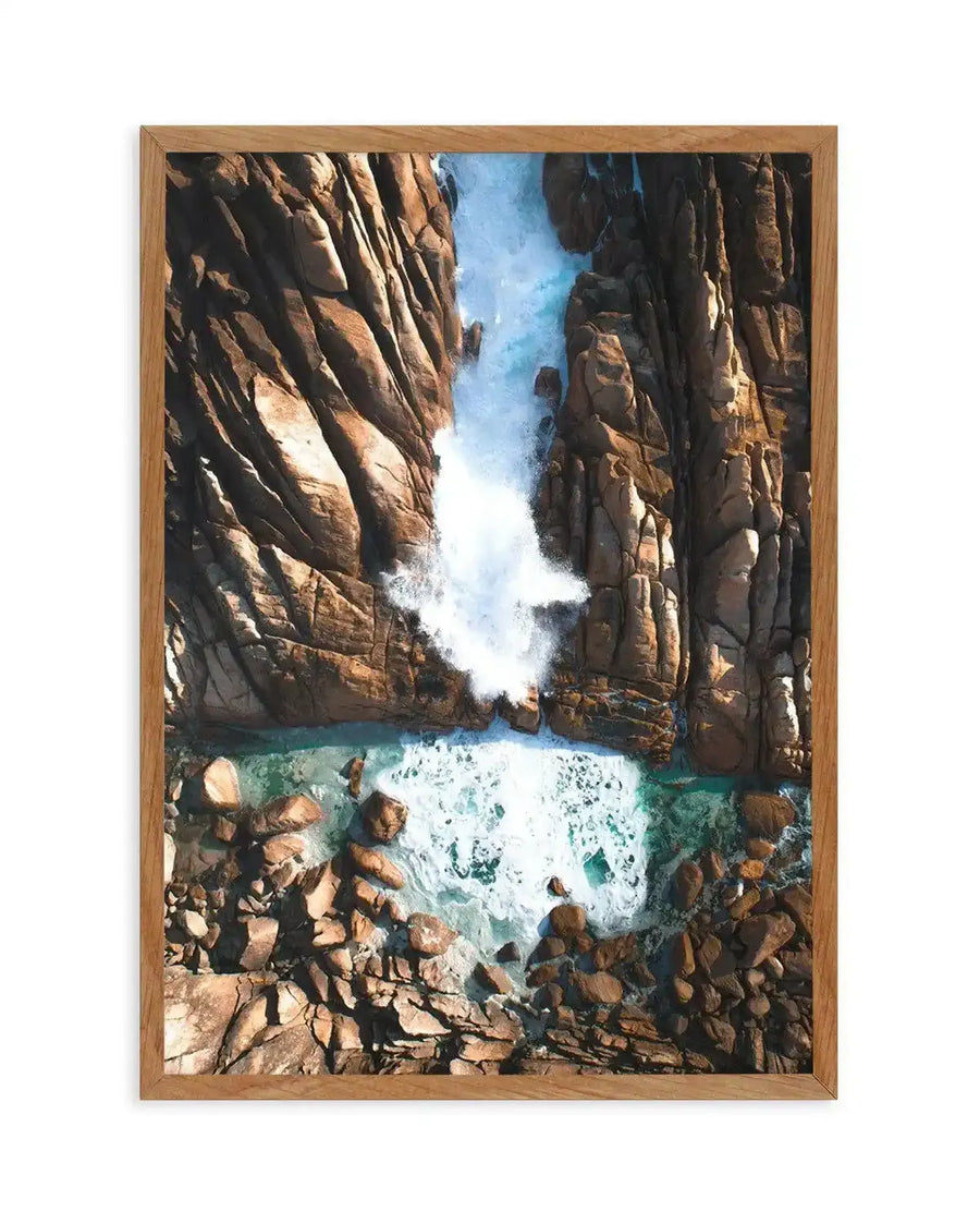 Framed aerial photograph of a natural spa with crashing waves against rugged brown rock formations, with a natural wood frame and white border. The composition captures the dynamic movement of white foamy water against the deep blue-green of the ocean and the warm tones of the rocky coastline.
