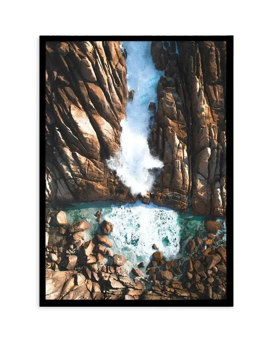Framed aerial photograph of Injidup Natural Spa in Western Australia, with a black frame and white border. The composition captures powerful turquoise waves crashing against rugged, reddish-brown rock formations, creating a dramatic spray of white foam. The clear, shallow water in the foreground reveals the rocky seabed.