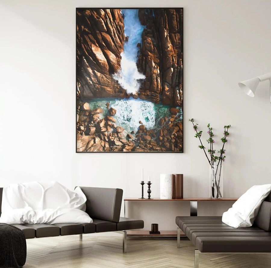 Lifestyle shot of a minimalist living room featuring a large framed art print of Injidup Natural Spa. The print, displayed on a white wall, captures an aerial view of turquoise ocean water crashing against rugged, warm brown rock formations, creating white foamy waves. The room includes two dark brown leather sofas with white cushions, a wooden console table with candles and a tall clear vase with flowering branches, and a white floor lamp.