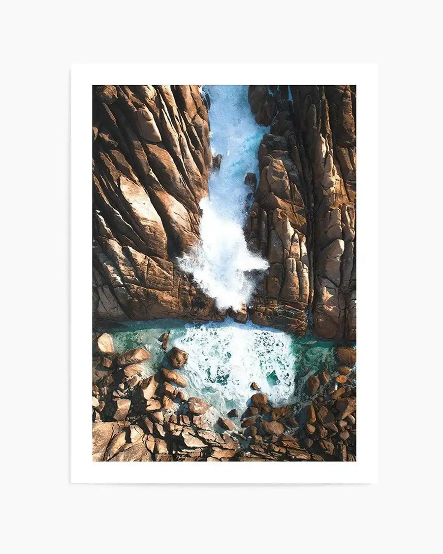 Framed aerial photography art print of Injidup Natural Spa in Western Australia, with natural wood frame and white border. The composition captures powerful turquoise waves crashing against rugged, sun-drenched brown rock formations, creating white foam and spray. The water settles into a calm, clear pool at the base of the rocks.
