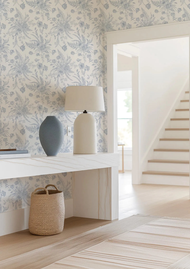 Lifestyle shot of a hallway featuring In the Hamptons wallpaper. The wallpaper has a repeating pattern of delicate blue floral and fern motifs on a cream background, creating a soft, botanical aesthetic. A white console table with a subtle marble pattern is against the wall, holding a textured blue vase and a cream-colored lamp. A woven basket sits on the floor beneath the table. In the background, a white staircase with light wood treads ascends, and a doorway reveals a bright room with a window.