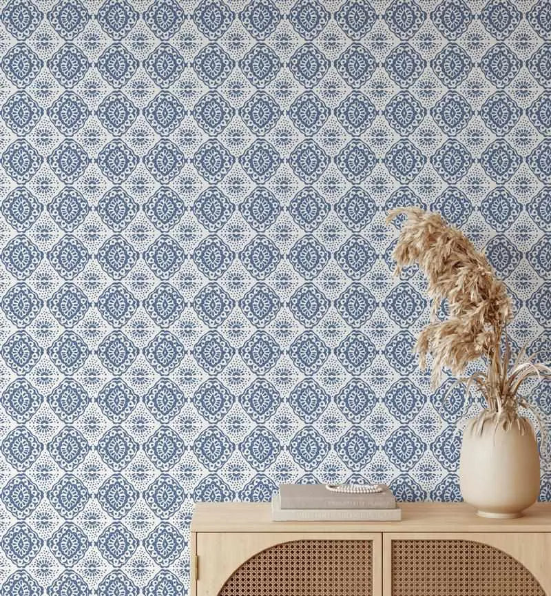 Ishka Wallpaper displayed on a wall above a light wood cabinet with woven rattan doors. The wallpaper features a repeating geometric damask pattern in teal blue and cream, with ornate diamond-shaped medallions containing intricate floral motifs. A cream ceramic vase with dried pampas grass and stacked books sit on the cabinet surface.