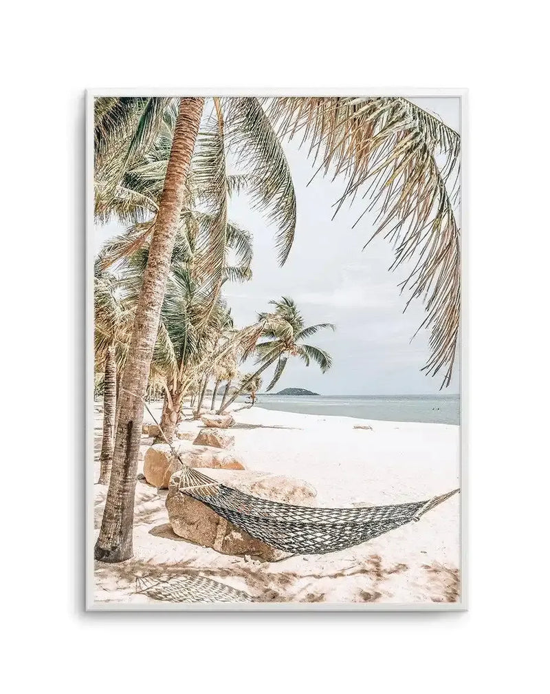 Framed photography art print of a tropical beach scene with a white border and natural wood frame. The image features a white sand beach with several tall palm trees on the left, and a woven black and white hammock strung between two large rocks in the foreground. The ocean is visible in the background under a pale blue sky, with a small island on the horizon.