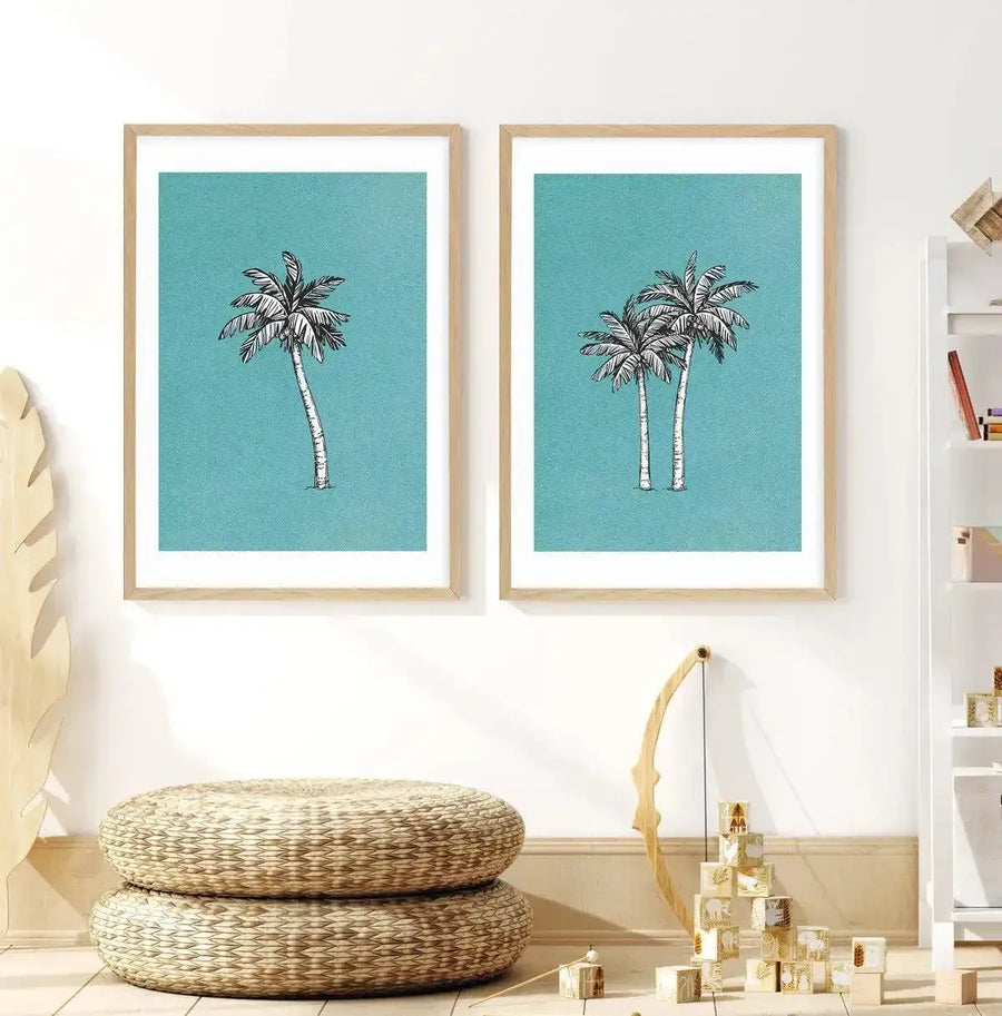 Kids Room:
Two framed art prints featuring palm trees displayed side-by-side on a white wall in a child's room. Each print depicts black and white illustrations of palm trees against a textured teal background, with natural wood frames and white borders. The left print shows a single palm tree, while the right print shows two palm trees. Below the prints are two stacked round woven floor cushions, a wooden toy bow, and a pyramid of wooden blocks with animal illustrations.