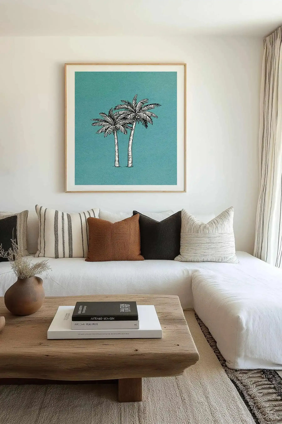 Lifestyle shot of a minimalist living room featuring a large framed art print of two palm trees with black and white fronds and trunks against a teal background, displayed above a white sectional sofa. The sofa is adorned with various textured throw pillows in black, white, and rust brown. A rustic wooden coffee table in the foreground holds a stack of books and a brown ceramic vase with dried grasses.