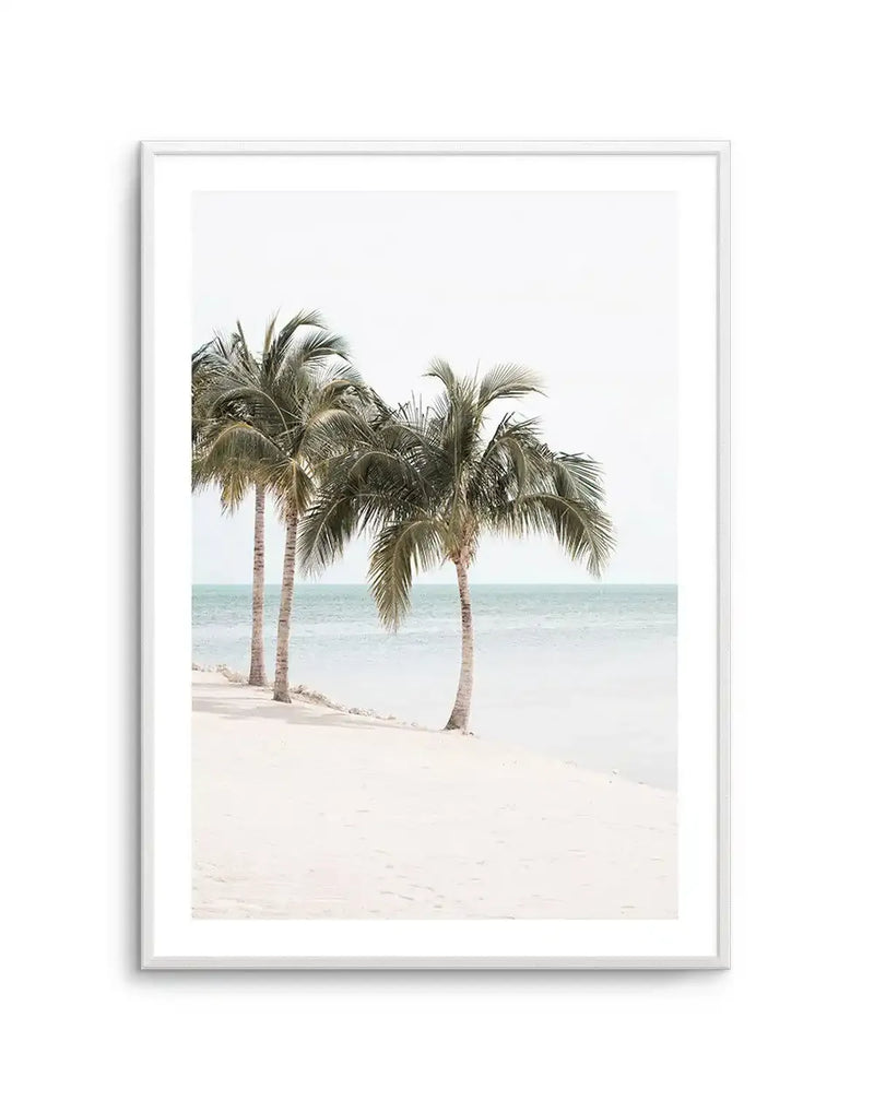 Framed photograph of a serene tropical beach scene with three palm trees on white sand, with a calm turquoise ocean and a bright white sky. The print is displayed in a white frame with a white border.