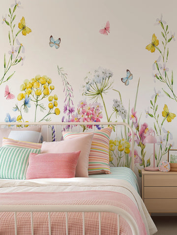 Peel and stick wallpaper mural displayed on a bedroom wall behind a white metal bed frame. The design features a watercolor-style botanical garden with delicate illustrations of yellow, pink, and white flowers including yarrow, sweet peas, and Queen Anne's lace on green stems scattered across a soft beige background. Blue and pink butterflies flutter throughout the composition. The mural coordinates with striped bedding in coral, green, and cream tones and pastel throw pillows on the bed below.