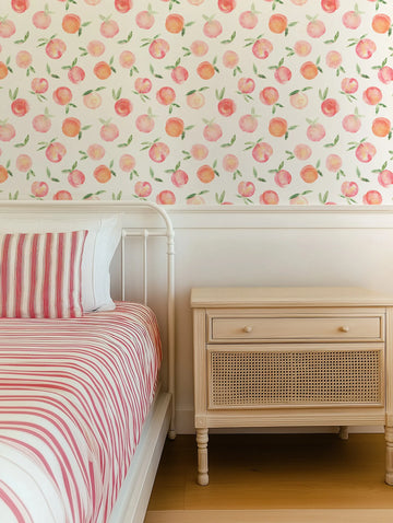 Lifestyle shot of a bedroom featuring a white metal bed frame with red and white striped bedding, and a light wood bedside table with a cane front drawer. The wall above the wainscoting is covered in wallpaper with a repeating pattern of hand-painted peaches in shades of pink and orange with green leaves, on a cream background.