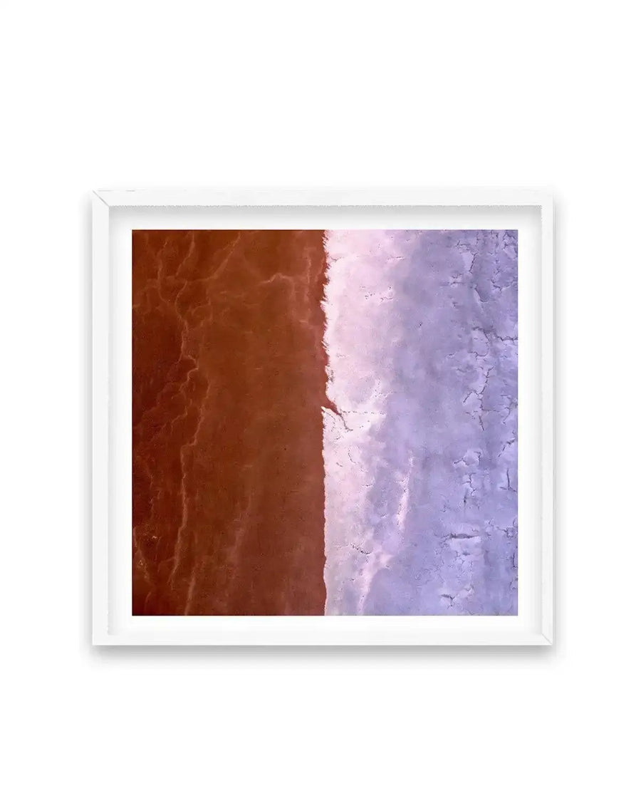 Framed aerial photography print of a vibrant landscape split vertically down the middle. The left side features rich, reddish-brown earth with intricate, dry cracked patterns, while the right side displays a contrasting pale lavender or light purple terrain with similar cracked textures, suggesting a dry lakebed or salt flats. The print is displayed in a white frame with a white border.