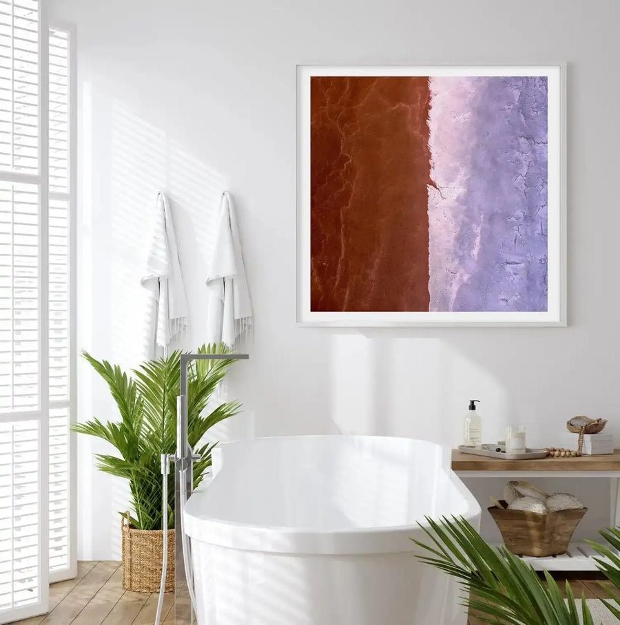 Large framed art print displayed above a white bathtub in a bright, minimalist bathroom. The print features an aerial photograph of a landscape divided vertically into two distinct halves: the left side is a rich, reddish-brown, while the right side is a pale, textured lavender-purple, separated by a jagged white line. The artwork is presented in a white frame with a white border. The bathroom includes a large potted palm plant, white towels, and a wooden shelf with bath accessories.