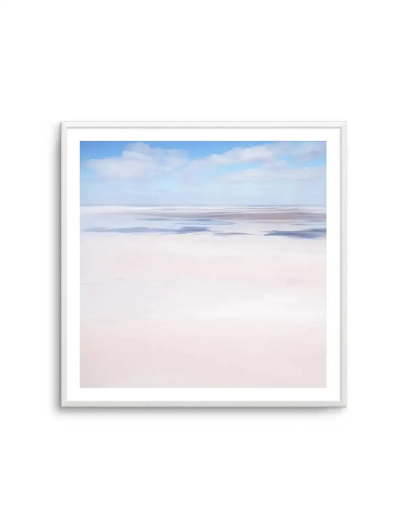 Framed photography art print of a vast, flat landscape under a blue sky with scattered white clouds, displayed in a white frame with a white border. The foreground is a pale, almost white, sandy or salt flat, transitioning to a horizon line where patches of darker, possibly wet or muddy ground, are visible. The overall impression is serene and expansive.