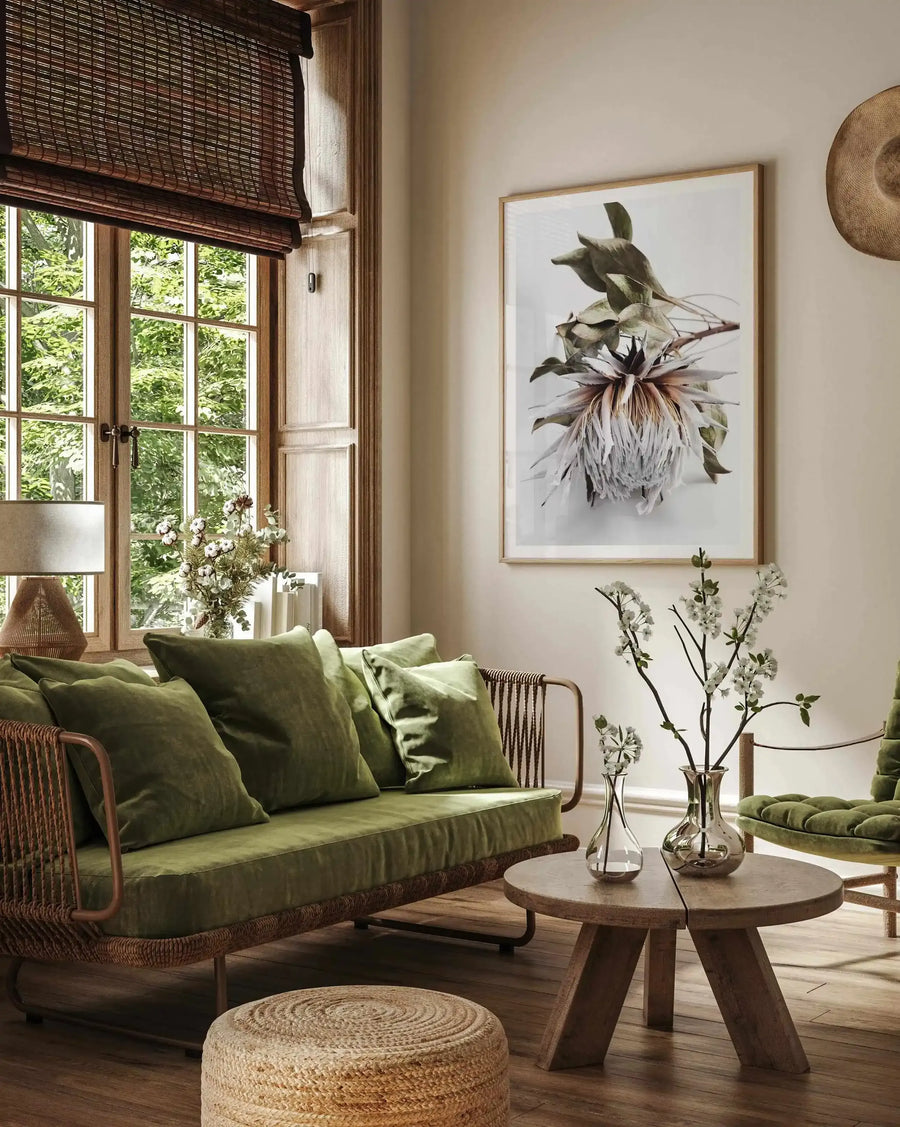 King Protea I Art Print displayed in a natural wood frame on a cream-colored wall. The botanical illustration depicts a large protea flower with pale pink and burgundy petals, dark green leaves, and delicate stamens, rendered in a soft watercolor style against a white background. The framed print is shown in a sunlit living room setting with a green upholstered sofa, woven rattan furniture, wooden side table with fresh flowers, and natural fiber accents.