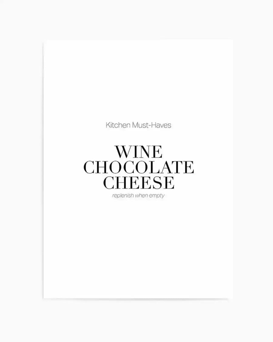 A minimalist art print featuring the text "Kitchen Must-Haves" in small gray letters at the top, followed by "WINE CHOCOLATE CHEESE" in large black serif capital letters in the center, with "replenish when empty" in small gray italicized text below. The design is set against a clean white background with a light gray vertical line on the left edge, creating a modern, typographic aesthetic with ample white space throughout.