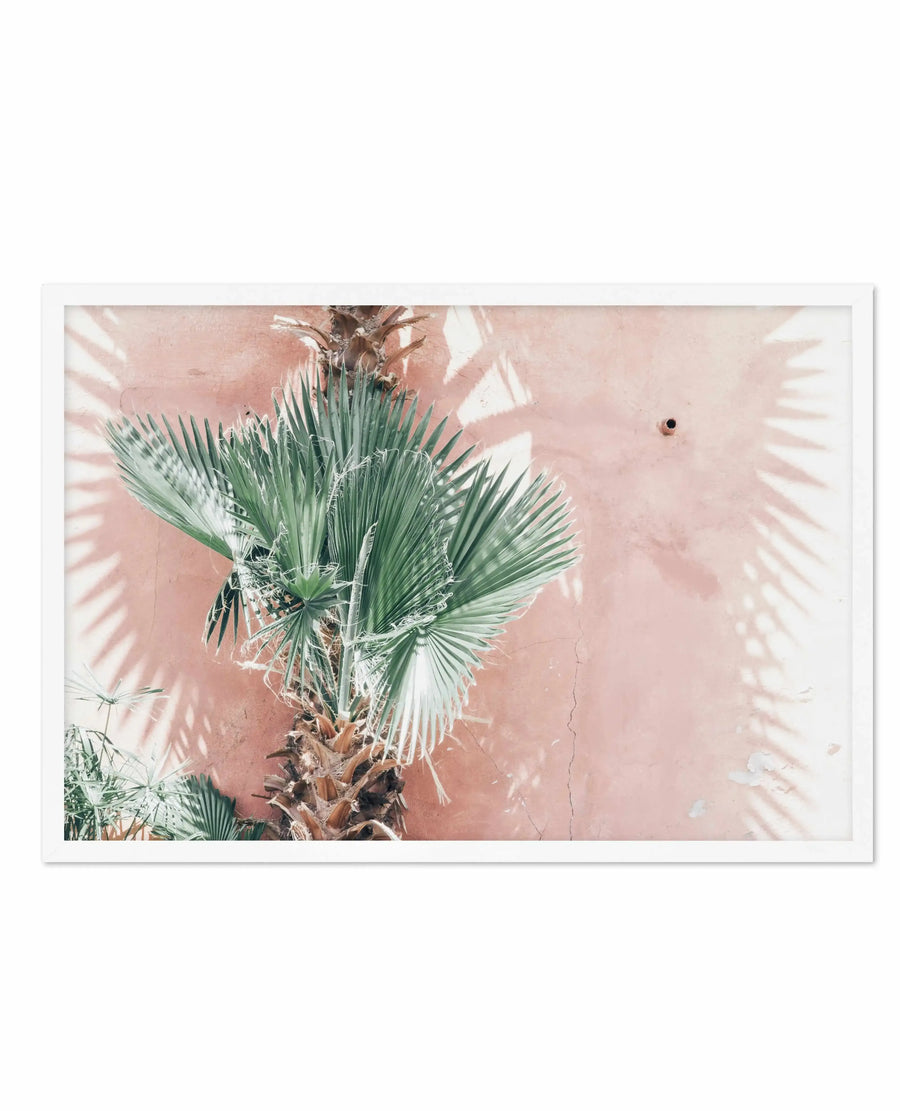 Overhead view of LA Palms art print displayed against a dusty rose-colored wall. The photograph features green fan palm fronds with sharp, radiating leaves casting linear shadows across the textured terracotta and cream wall surface. A small dark object visible in the upper right. The print is shown with a white border frame, capturing a minimalist, botanical aesthetic with warm earthy tones and natural lighting creating a serene, Mediterranean mood.