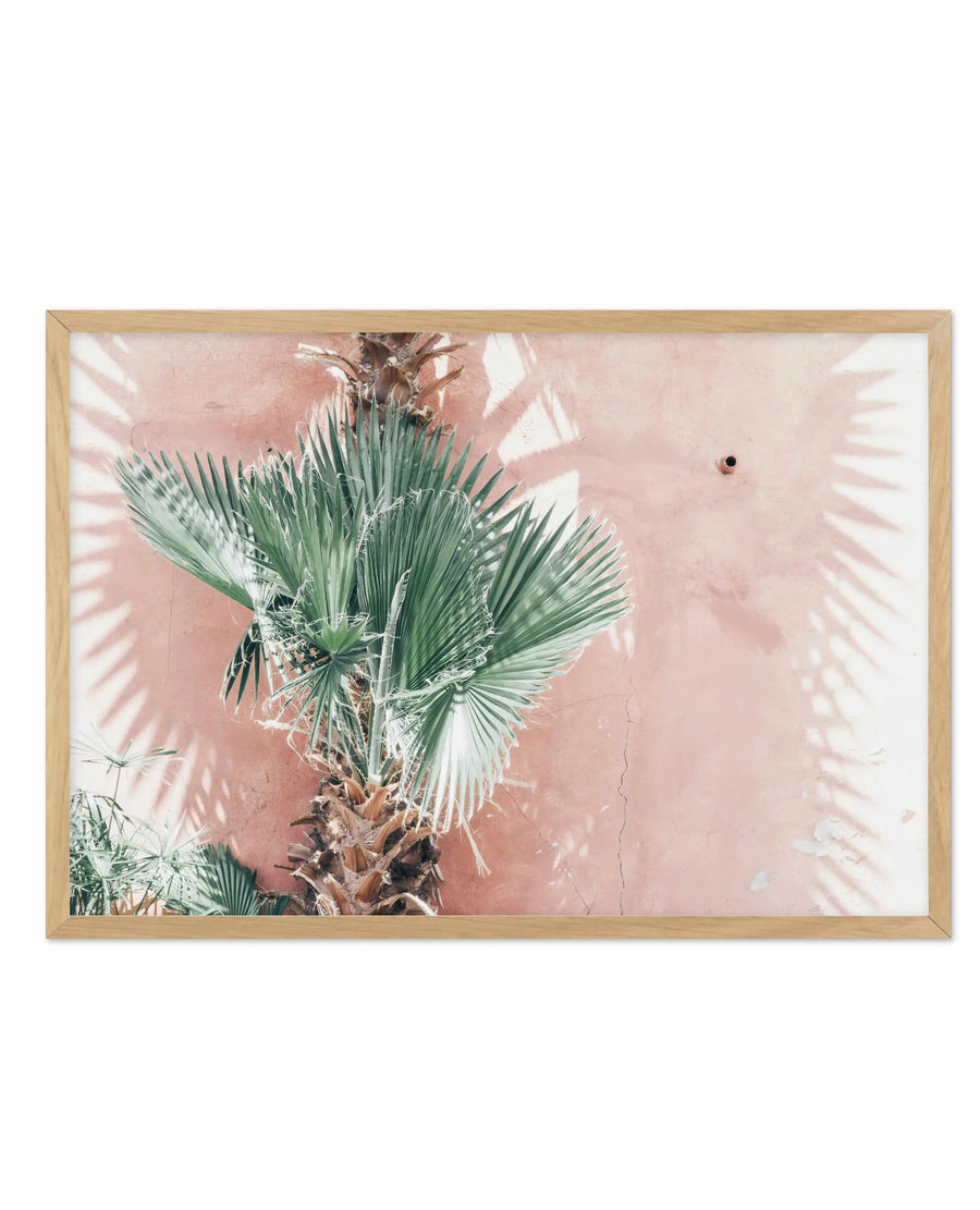 LA Palms art print in natural wood frame featuring a photograph of fan palm fronds with green strappy leaves against a dusty rose pink wall. Bright sunlight casts sharp linear shadows across the wall and foliage. A smaller palm plant is visible in the lower left corner. The photograph has a warm, tropical aesthetic with high contrast between the vivid green leaves and the soft muted pink background. White border surrounds the image within the frame.