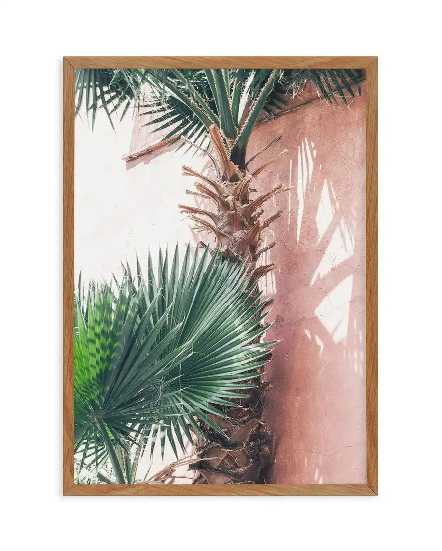 Framed art print featuring a close-up botanical composition of palm fronds against a terracotta-colored wall. The artwork displays vibrant green feathered palm leaves in the foreground and softer, darker green foliage at the top, with warm peachy-brown tones of the palm trunk visible throughout. The style appears to be watercolor or mixed media with soft, painterly qualities. The print is mounted in a natural wood frame with a white border, creating a warm, tropical aesthetic.