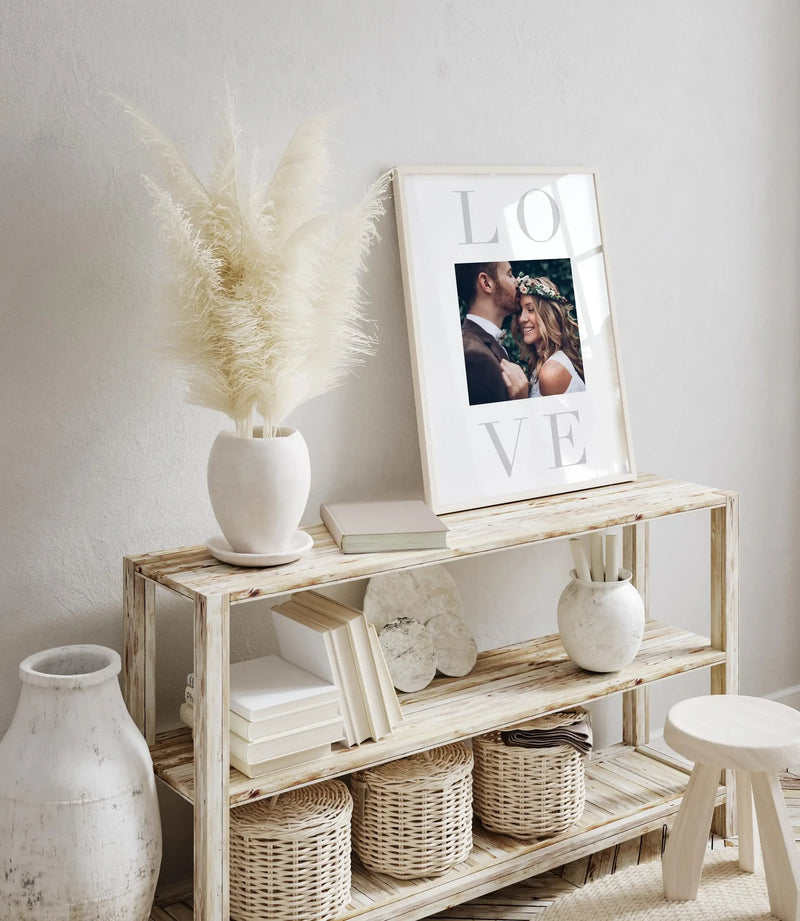 LOVE custom photo art print displayed in a white frame with white border on a light wood shelf. The print features a photograph of a couple in the center, surrounded by large gray letters spelling "LOVE" vertically. The frame leans against a white wall above a wooden shelving unit styled with cream pampas grass, white ceramic vases, stacked books, woven baskets, and a small white stool, creating a minimalist, neutral-toned home décor scene.