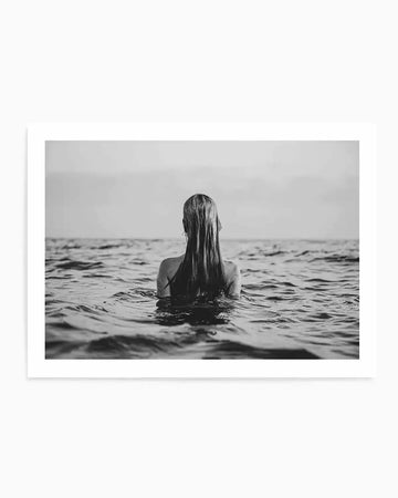 Framed black and white photograph of a woman standing in the ocean with her back to the viewer, with a natural wood frame and white border. Her long, wet hair hangs down her back, and the water reaches her chest, creating gentle ripples around her. The horizon line is visible in the distance under an overcast sky.
