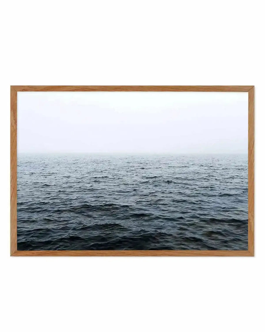 Framed photograph of a vast, calm ocean under a misty, overcast sky, printed on fine art paper and displayed in a natural oak frame with white border. The water transitions from dark, deep blue in the foreground to a lighter grey-blue towards the horizon, where it blends seamlessly with the pale, hazy sky.