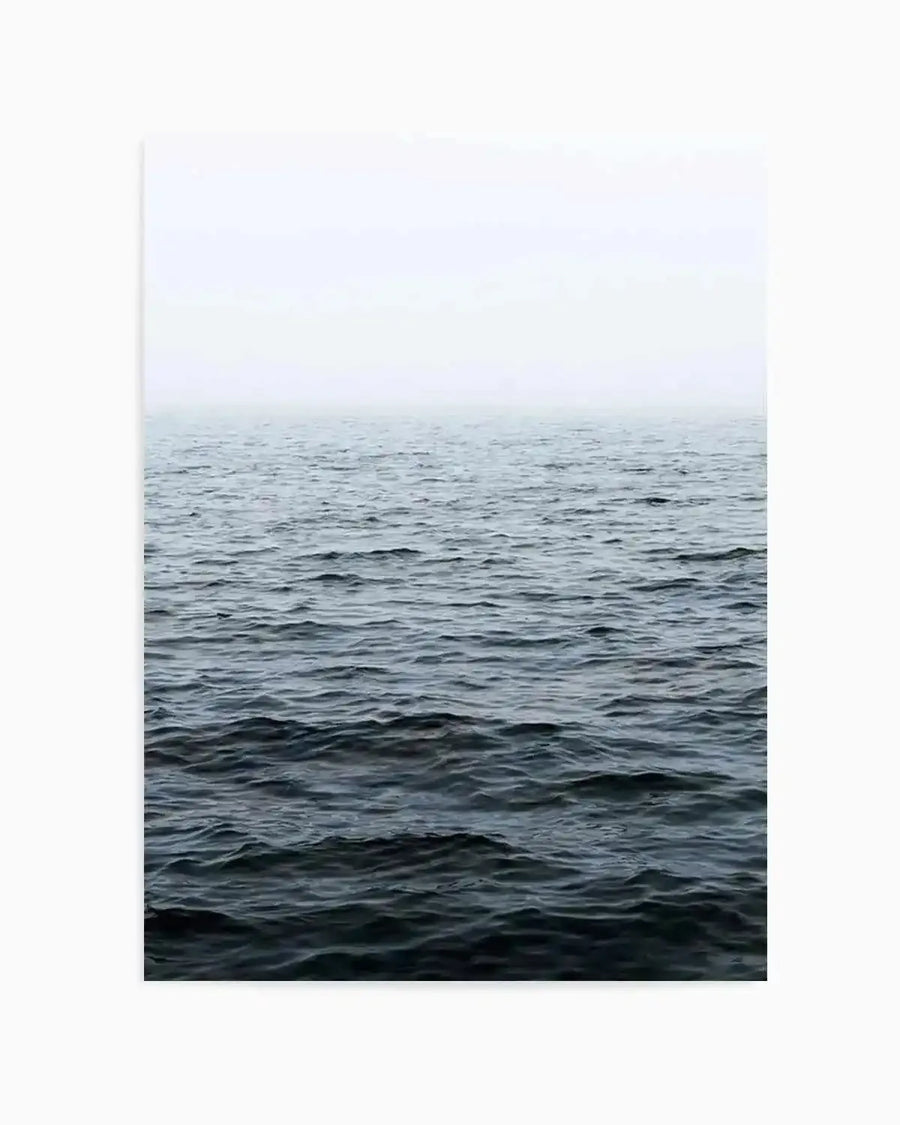"Photography art print featuring a minimalist ocean scene with a calm, dark blue-grey sea under a hazy, light grey sky. The horizon line is subtly defined, creating a serene and expansive mood with gentle ripples visible on the water's surface."