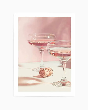 Photograph of two coupe champagne glasses filled with sparkling rosé wine, with a champagne cork lying on a white surface in the foreground. The background is a soft, diffused blush pink, creating a dreamy and celebratory mood. The glasses reflect light, highlighting the effervescence of the wine.