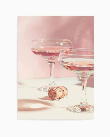 Photography art print featuring two coupe glasses filled with sparkling rosé champagne, with a champagne cork and wire cage lying on a white surface in the foreground. The background is a soft, diffused blush pink, creating a celebratory and elegant mood.