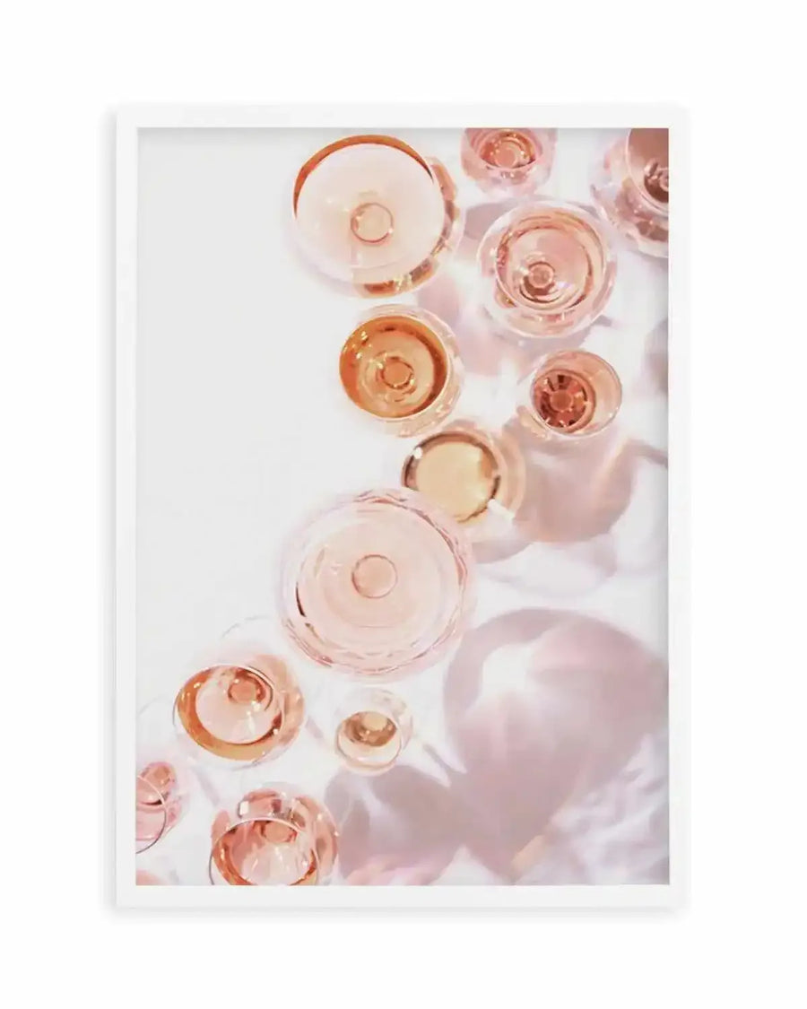 Framed art print featuring an overhead photograph of multiple glasses filled with rosé champagne, casting soft pink shadows on a white surface, with a white frame and white border. The composition shows glasses of varying sizes and shapes, some with a golden hue, arranged organically across the frame.